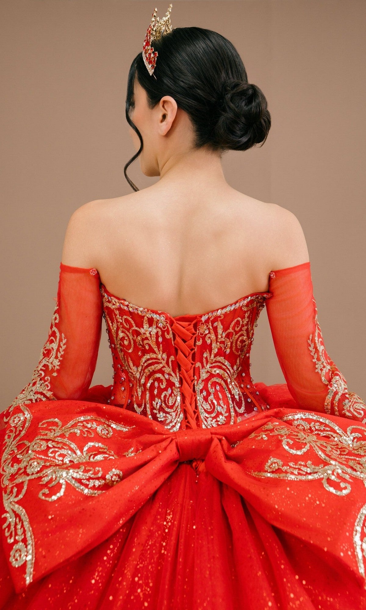 A woman seen from behind wears the Quinceanera Dress 8545JQ By Cinderella Couture, featuring gold embroidery, a large bow, and an off-the-shoulder sweetheart neckline; her hair is in a low bun with delicate adornments.
